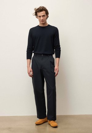 Tânăr stând în picioare în fața unui perete simplu, purtând o cămașă neagră cu mâneci lungi, pantaloni negri și pantofi tip loafer maro deschis.