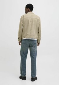 Beige Wildlederjacke mit geradem Schnitt, Gesäßtaschen und geknöpften Ärmeln, getragen über blauen lässigen Jeans und schwarzen Schuhen.