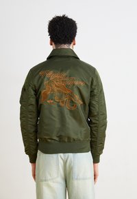 Olivno zelena bomber jakna z vezenim tigrom in napisom "Flying Tigers" na hrbtu, z rebrastimi manšetami in pasom.