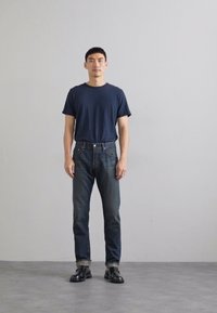 Dunkelblaues Baumwoll-T-Shirt, schmal geschnittene dunkle Denim-Jeans und schwarze Lederschuhe. Das Modell steht vor einem hellgrauen Hintergrund.