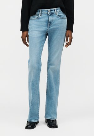 Άτομο που φοράει ανοιχτό μπλε τζιν με ίσια γραμμή, μαύρο πουλόβερ και μαύρα παπούτσια με τετράγωνη μύτη, στέκεται μπροστά σε απλό λευκό φόντο.