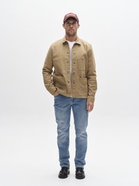 Smėlio spalvos drobinė striukė su sagomis, keturios priekinės kišenės ir aplikacija. Dėvima virš baltos marškinės ir derinama su mėlynais džinsais bei juodais batais.