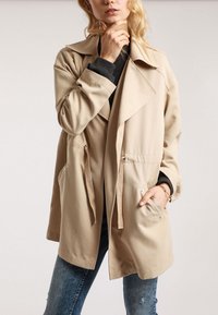 Beige trenssitakki leveällä kauluksella, sivutaskuilla ja vyötärön kiristysnauhalla. Sileä kangas ja hieman yläkanttiin oleva istuvuus.