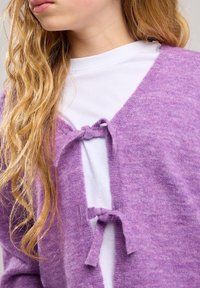 Cárdigan de punto morado con escote en V, lazos frontales y una textura suave. Usado sobre una camisa blanca, que resalta su delicada confección.