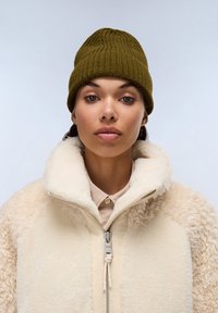 Gorro de punto en color oliva combinado con una chaqueta cremosa con cremallera y forro de sherpa; presenta una textura suave y un cuello elevado; el cabello es parcialmente visible.