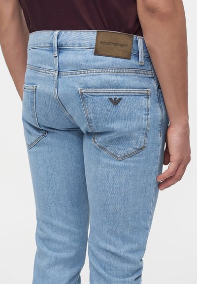 Άνδρας που φοράει ανοιχτό μπλε τζιν Emporio Armani με λογότυπο στη ζώνη και μεταλλικό λογότυπο στην δεξιά πίσω τσέπη, στέκεται μπροστά σε μονόχρωμο φόντο.