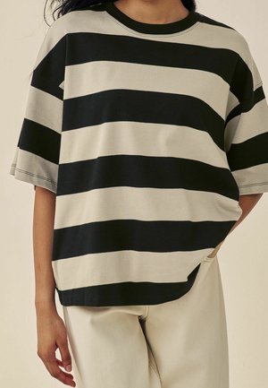 Person iført overdimensioneret kortærmet skjorte med brede sorte og beige vandrette striber, kombineret med lysebeige bukser.