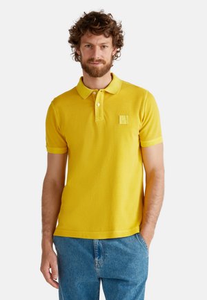 Camisa polo amarilla de manga corta, con un cuello clásico y un pequeño parche de logo en el pecho. La tela parece suave y ligera.
