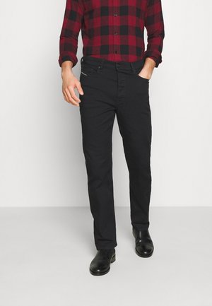 Man draagt zwarte slim-fit broek, zwarte schoenen en een rood-zwarte geruite overhemd met lange mouwen, één hand in de zak.