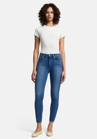Blå denim skinny jeans med hög midja, kombinerade med en figurnära vit bomulls-T-shirt och ljusfärgade platta skor. Lockigt hår syns.