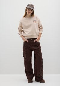 Felpa beige con grafica "NATALE" e alberi, abbinata a pantaloni cargo marroni con molteplici tasche e vestibilità comoda. Berretto grigio completa il look.