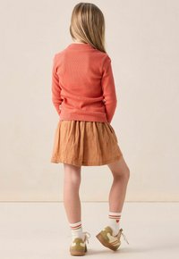 Koraalkleurig ribgebreid langmouwtopje gecombineerd met een roestkleurige rok met bloemmotief; wit en rood gestreepte sokken; beige en groene sneakers.