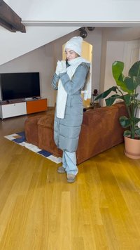 Lang, lyseblå pufferjakke med hætte, parret med et hvidt tørklæde, handsker og hat. Beige slippers, trægulv og en plante i baggrunden.