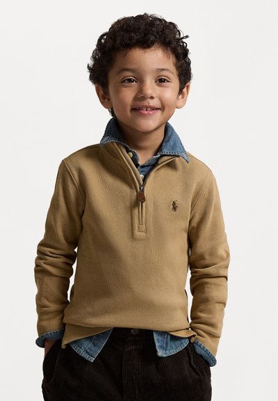 Smiling young boy with curly hair wears tan quarter-zip sweater over a blue denim shirt, hands in dark corduroy pants pockets, against white background.