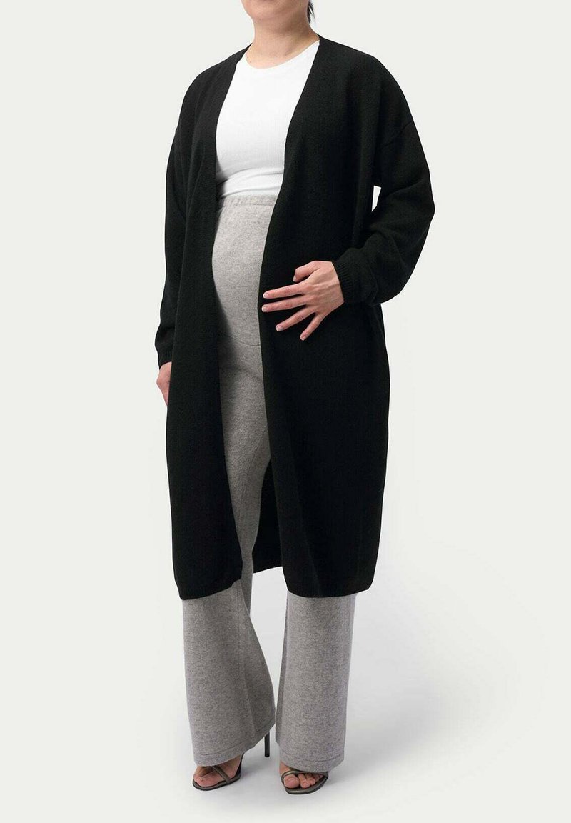 Svart cardigan med öppen front, långa ärmar och avslappnad passform, bärs över en vit topp och grå vida byxor. Slät textur.