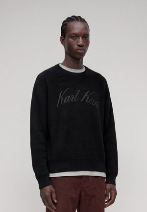 Jeune homme aux cheveux tressés portant un pull noir Karl Kani par-dessus une chemise blanche et un pantalon marron sur un fond clair uni.