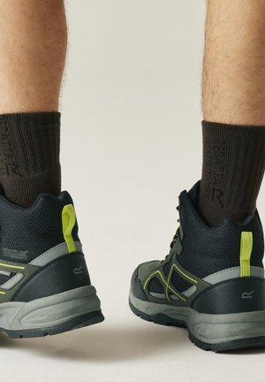 Nogi w ciemnych skarpetkach i czarnych, outdoorowych butach turystycznych z szarymi podeszwami i żółtymi pętelkami do ściągania, stojące na jasnej powierzchni.