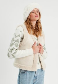 Beige Fleece-Weste mit cremefarbenem Schafslederbesatz, getragen über einem gemusterten hellen Pullover. Das Model trägt einen flauschigen weißen Hut. Lockere Jeans aus Denim sind sichtbar.