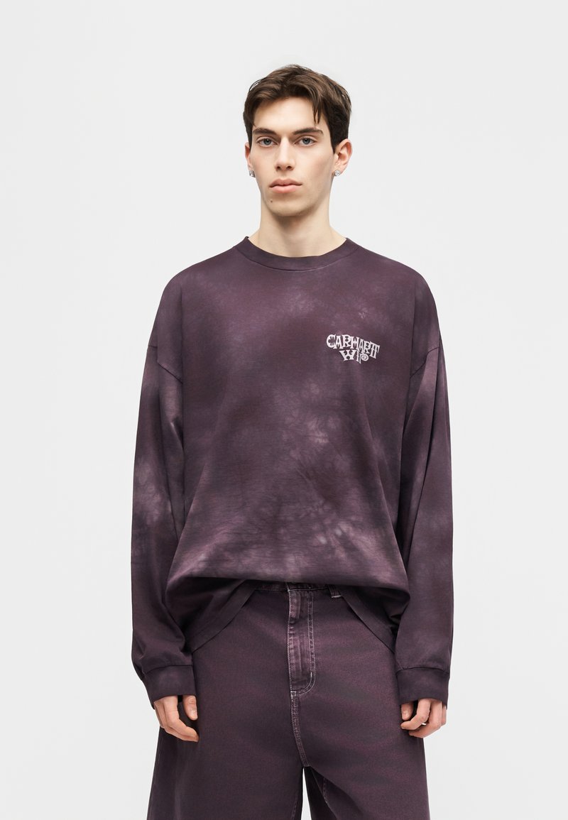 Giovane uomo che indossa una felpa larga tie-dye viola scuro con il logo "Carhartt" sul petto e pantaloni ampi abbinati, in piedi davanti a uno sfondo semplice.