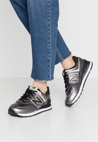 Scarpe da ginnastica nere e grigio metallizzato con superficie testurizzata, caratterizzate da un importante logo "N" argentato e lacci neri, montate su una suola bianca.