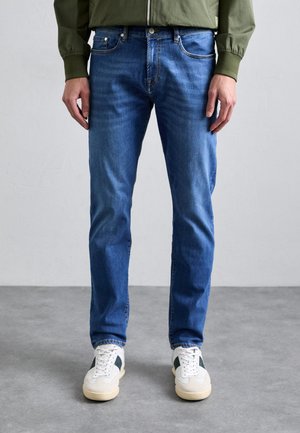 Blue denim jeans with a slim fit, featuring classic pockets and light fading. Paired with white sneakers with green accents.