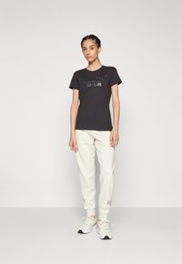 Camiseta negra con un diseño gráfico, combinada con pantalones de chándal color crema y zapatillas ligeras. Ropa deportiva simple y casual.