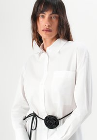 White button-up shirt with a breast pocket, worn with a black floral belt. Smooth fabric, loose fit, long sleeves with button cuffs.