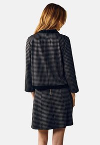 Sæt bestående af jakke og nederdel i sort og hvid ternet houndstooth-mønster, med trekvartærmer og en glat, tekstureret overflade, samt lynlåsdetalje på bagsiden.