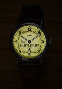 Montre-bracelet ronde Timex avec cadran jaune, chiffres noirs, texte Hufflepuff et symboles de la maison, aiguilles des heures et des minutes noires, et bracelet en cuir noir.
