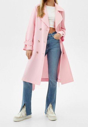 Trench-coat rose clair avec un design à double boutonnage, grand col et manches retroussées, associé à un jean bleu avec des fentes sur les côtés et des baskets blanches.