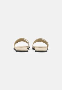 Paire de sandales plates beiges avec semelles noires, vues de dos, sur un fond gris clair uni.