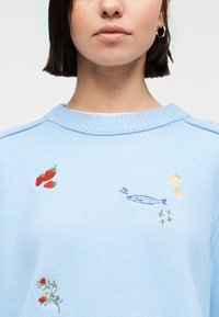 Γυναίκα που φοράει ανοιχτό μπλε πλεκτό πουλόβερ με κεντημένα φράουλες, ψάρια, φέτες λεμονιού και μικρά μοτίβα με άνθη και ελιά.