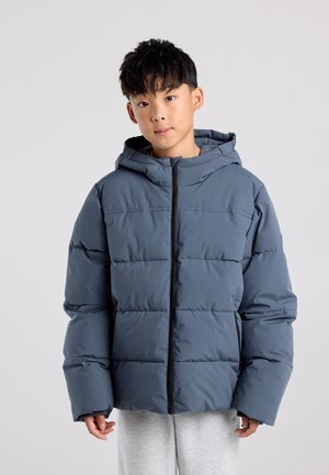 Jongen draagt een blauwe donsjas met capuchon en grijze joggingbroek, staand tegen een effen lichte achtergrond.
