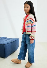 Colorful knitted cardigan with multicolored stripes, red top, wide blue jeans, and tan slip-on shoes, set against a neutral background.