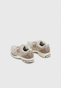 Ett par beiga och bruna New Balance löparskor med detaljer av mesh och mocka på vit bakgrund, sedda från bakre vänstra sidan.