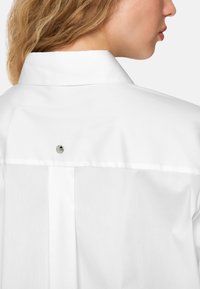 White shirt with a structured collar, featuring a metallic button detail at the back, and smooth, slightly textured fabric.