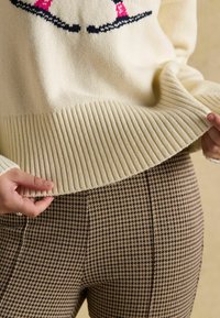 Cremfarbener, gerippter Strickpullover mit grafischem Muster, kombiniert mit braunen Hahnentritt-Musterleggings. Schwerpunkt auf Texturen und Farben.