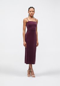 Femme aux cheveux tressés portant une robe midi ajustée sans manches de couleur violette et des chaussures blanches à bride à la cheville, debout devant un fond clair uni.