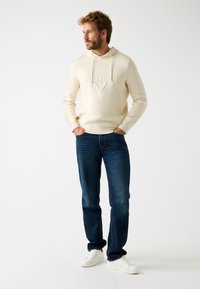 Sudadera con capucha color crema con el logo "GUESS" en relieve, combinada con jeans azul oscuro y zapatillas blancas, de pie frente a un fondo claro.