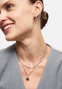 Silberne Ohrringe und geschichtete Halsketten. Eine Halskette hat ein glattes, geschwungenes Design, während die andere einen kleinen Elefantenanhänger trägt.