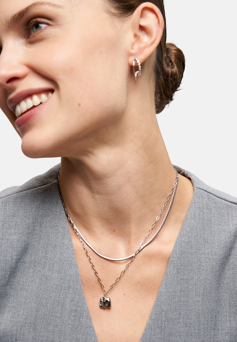 Silberne Ohrringe und geschichtete Halsketten. Eine Halskette hat ein glattes, geschwungenes Design, während die andere einen kleinen Elefantenanhänger trägt.