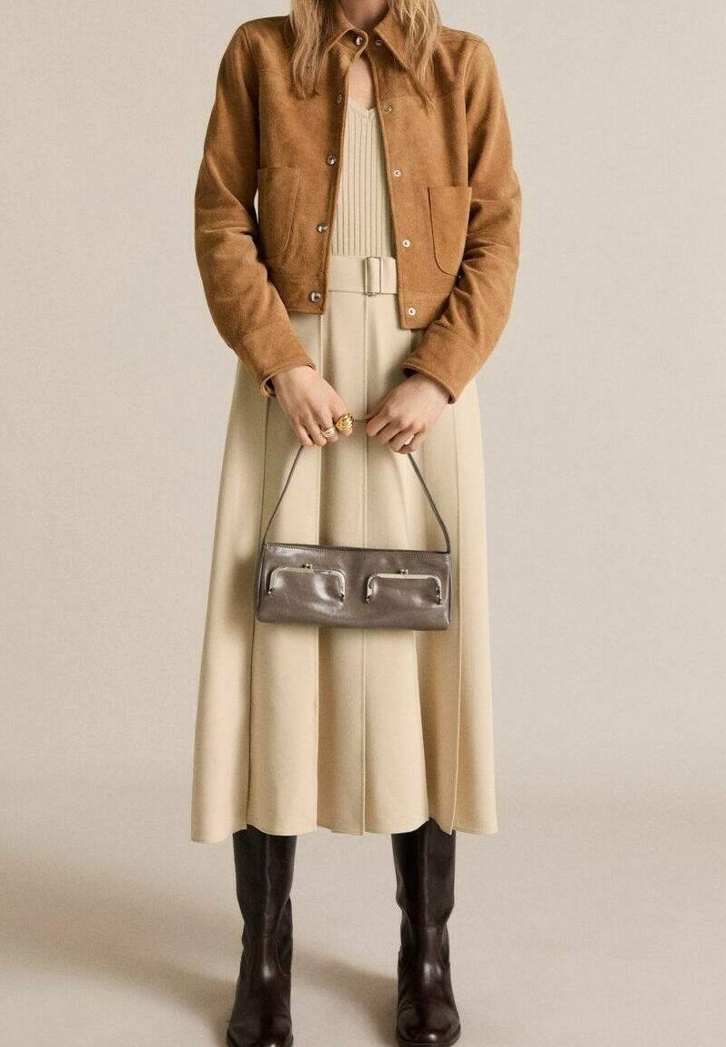 Brown suede jacket with snap buttons and two front pockets, paired with a cream pleated dress and knee-high brown leather boots. Gray handbag included.