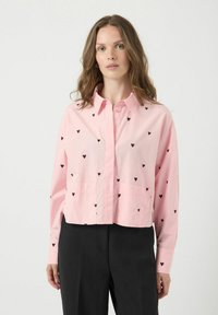 Chemise rose courte avec motifs de cœurs noirs, boutonnière devant, col et deux poches avant ; associée à un pantalon noir en coton fin.