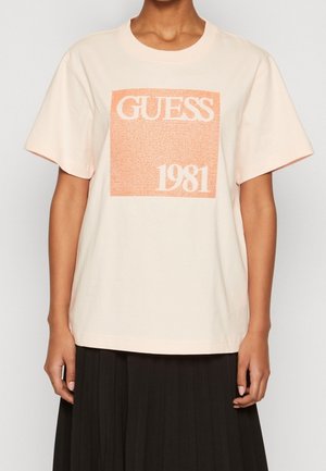 Personne portant un t-shirt beige avec une impression carrée orange indiquant « GUESS 1981 », associé à une jupe plissée noire.