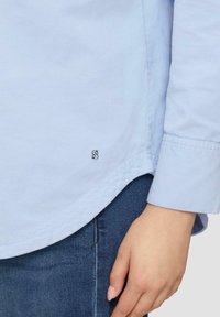 Lichtblauwe shirt gemaakt van zachte stof, met een subtiele textuur en een klein logo-detail nabij de zoom. Lange mouwen en een gebogen zoomontwerp.