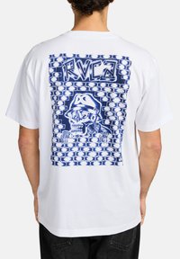 White cotton t-shirt featuring a blue graphic print of a skull wearing a hat, set against a brick wall design. Relaxed fit, short sleeves.