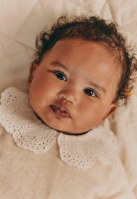 Krøllet hår baby med mørke øjne iført et beige outfit med en hvid blondekrave, liggende på et lyst farvet tæppe.