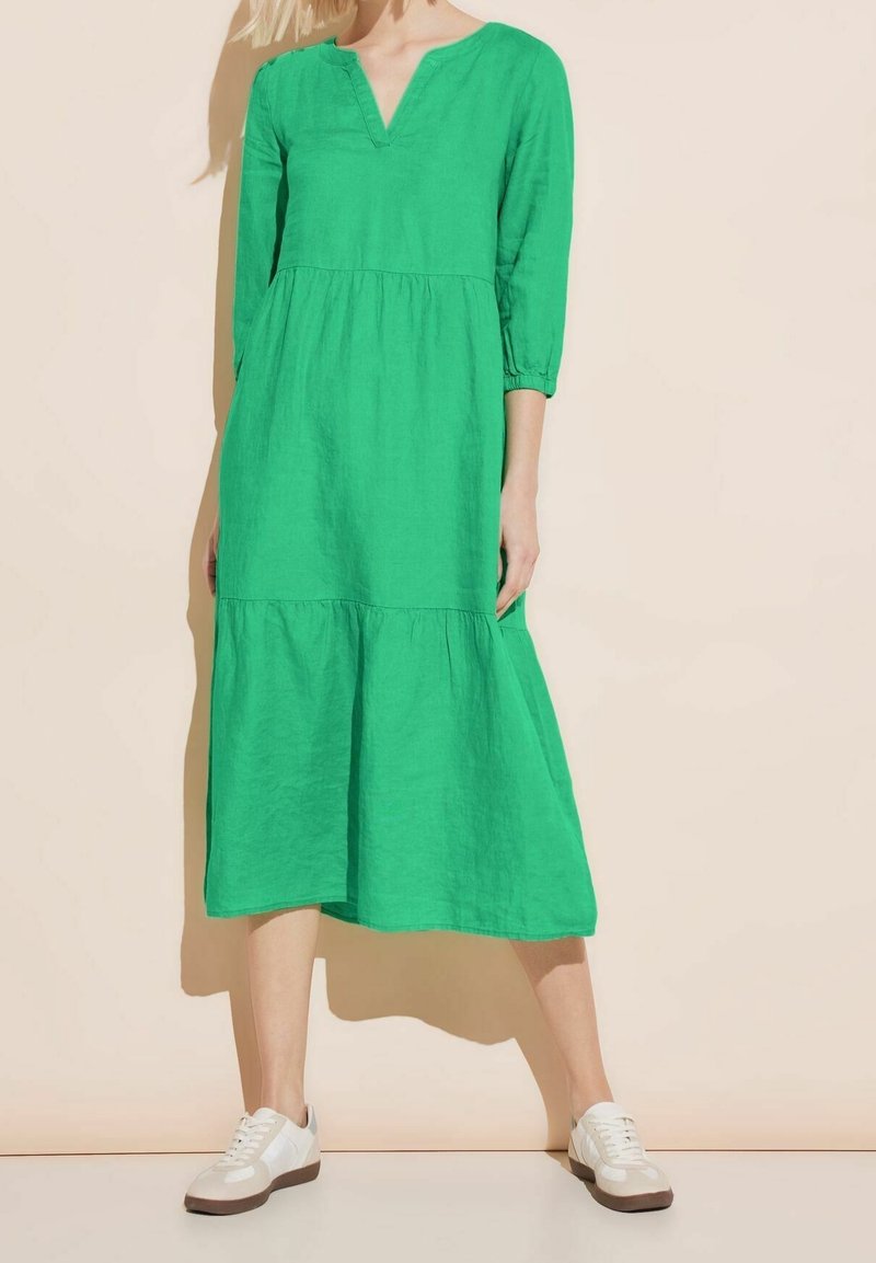 Femme vêtue d'une robe midi en lin vert vif à manches longues associée à des baskets blanches décontractées, debout devant un fond beige.