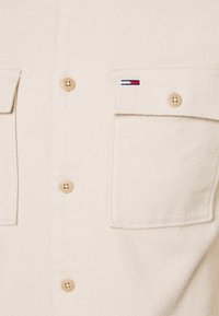 Primo piano di una camicia beige con bottoni, dotata di due tasche frontali; una flap della tasca presenta un piccolo logo rettangolare rosso, bianco e nero.