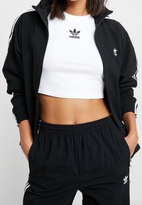 Černá tepláková souprava s mikinou na zip s bílými pruhy a logem, doplněná bílým krátkým tričkem a černými kalhotami s elastickým pasem.